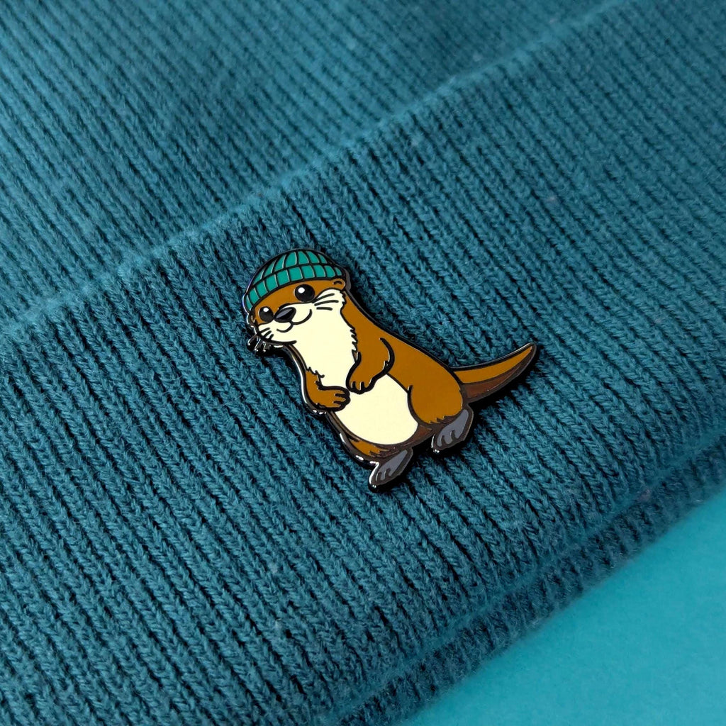 Pin of an otter wearing a green beanie on a textured blue surface