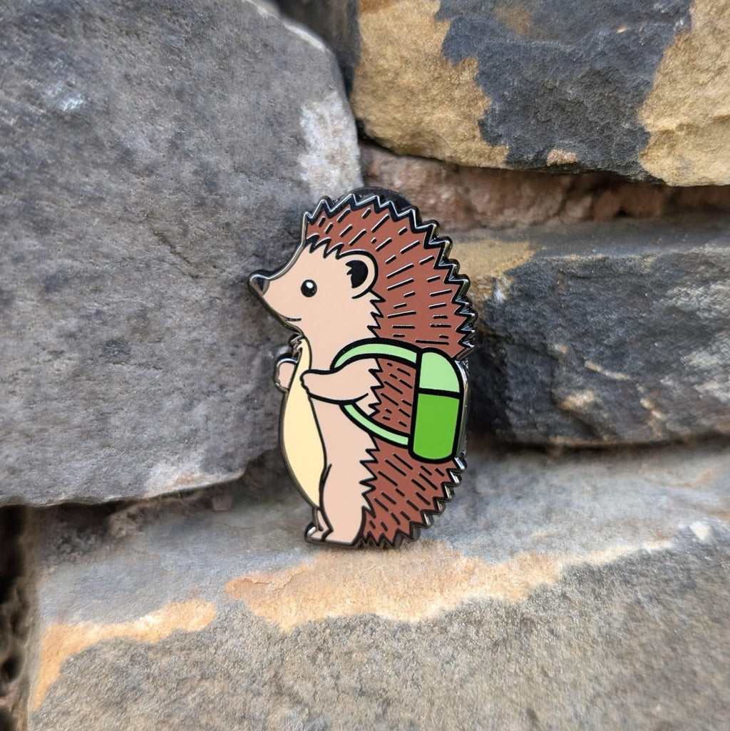 Hedgehog enamel pin with a green backpack on a stone surface