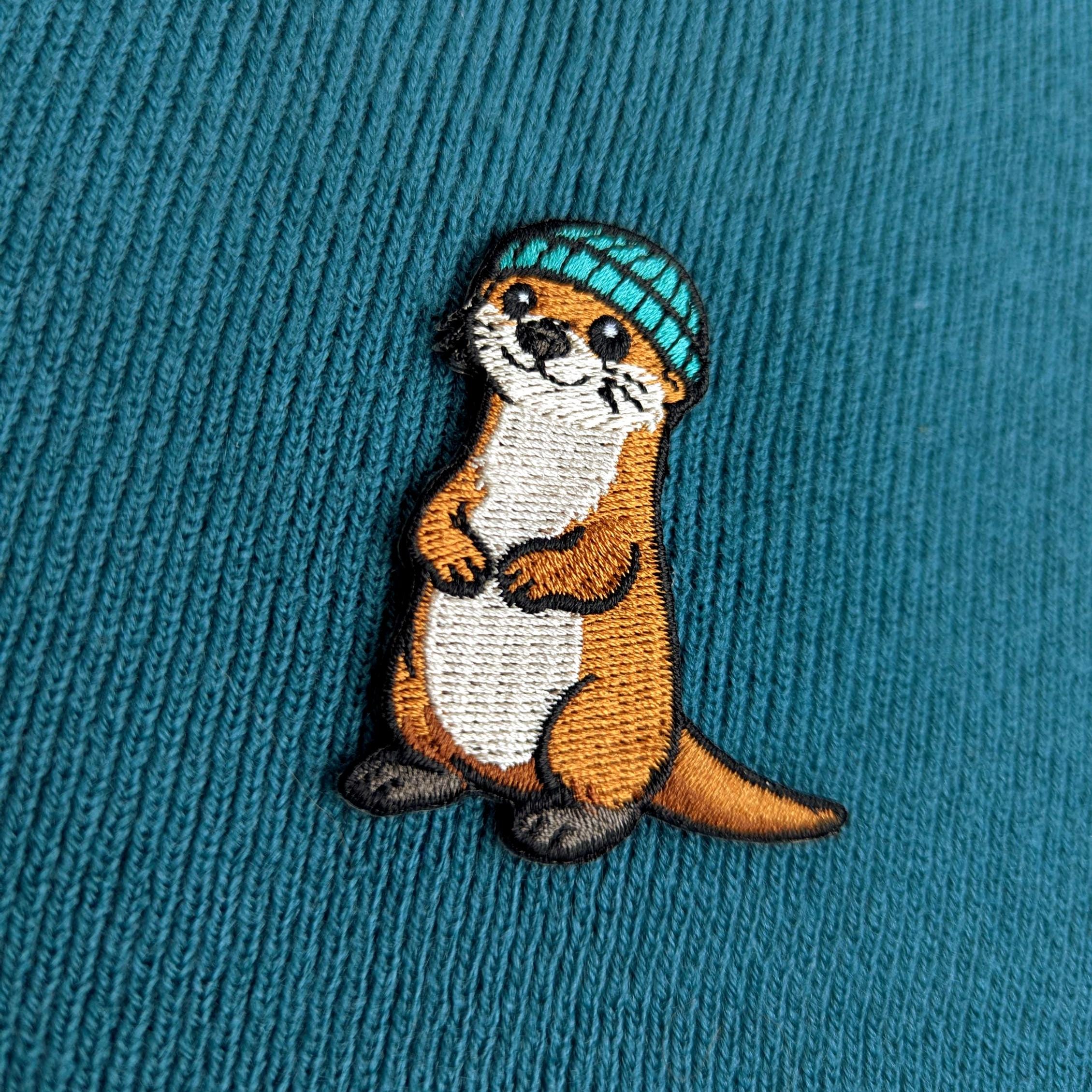 Embroidered otter wearing a blue hat on a teal fabric background