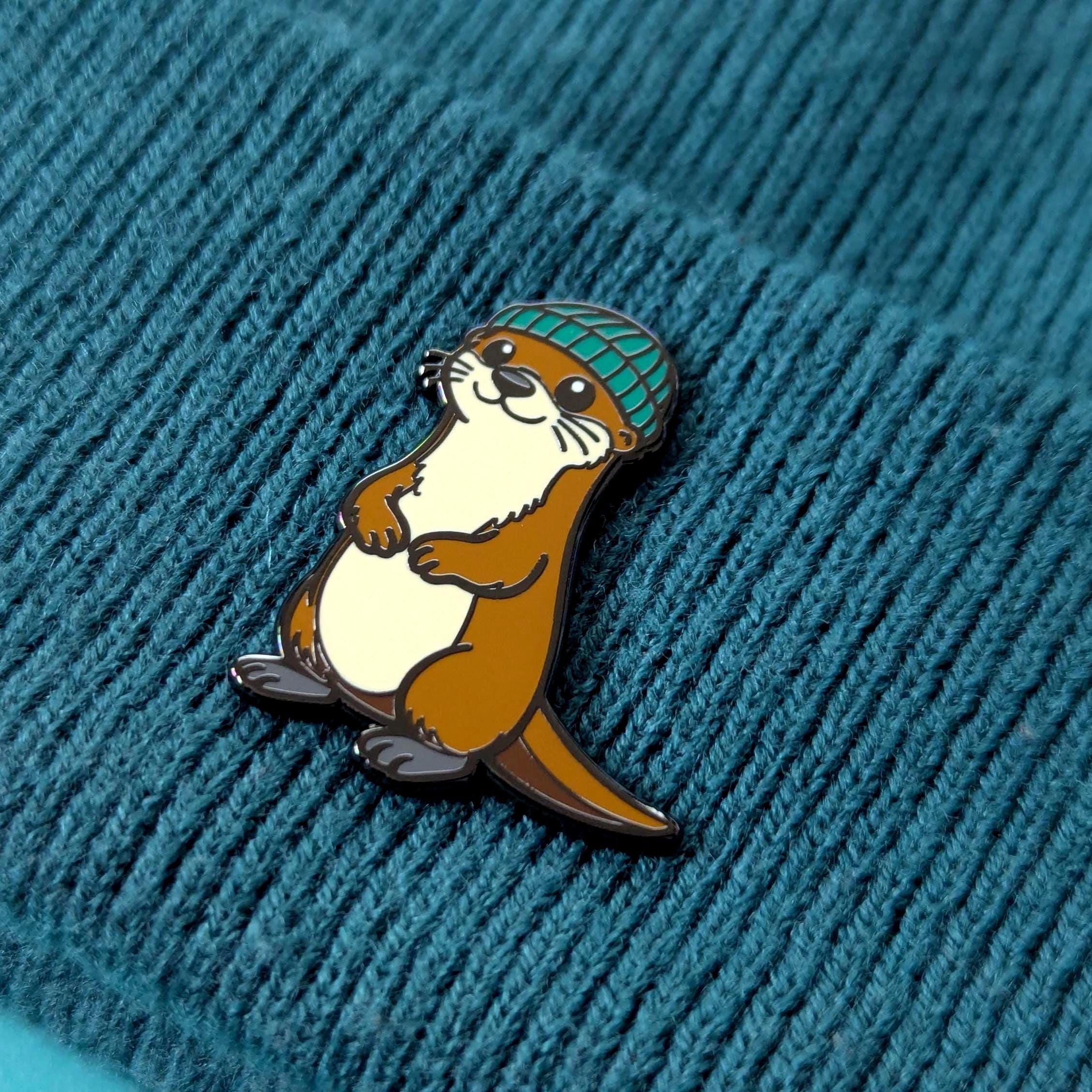 Pin of an otter wearing a green beanie on a textured blue background