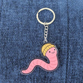 Keychain with a pink worm design wearing a yellow hat on a denim background
