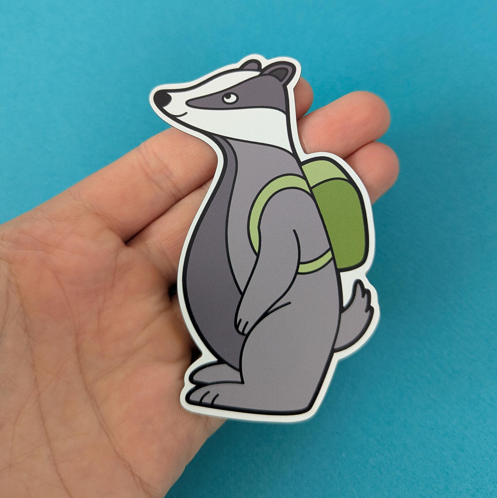 Hand holding a die cut magnet of a badger with a green backpack against a blue background