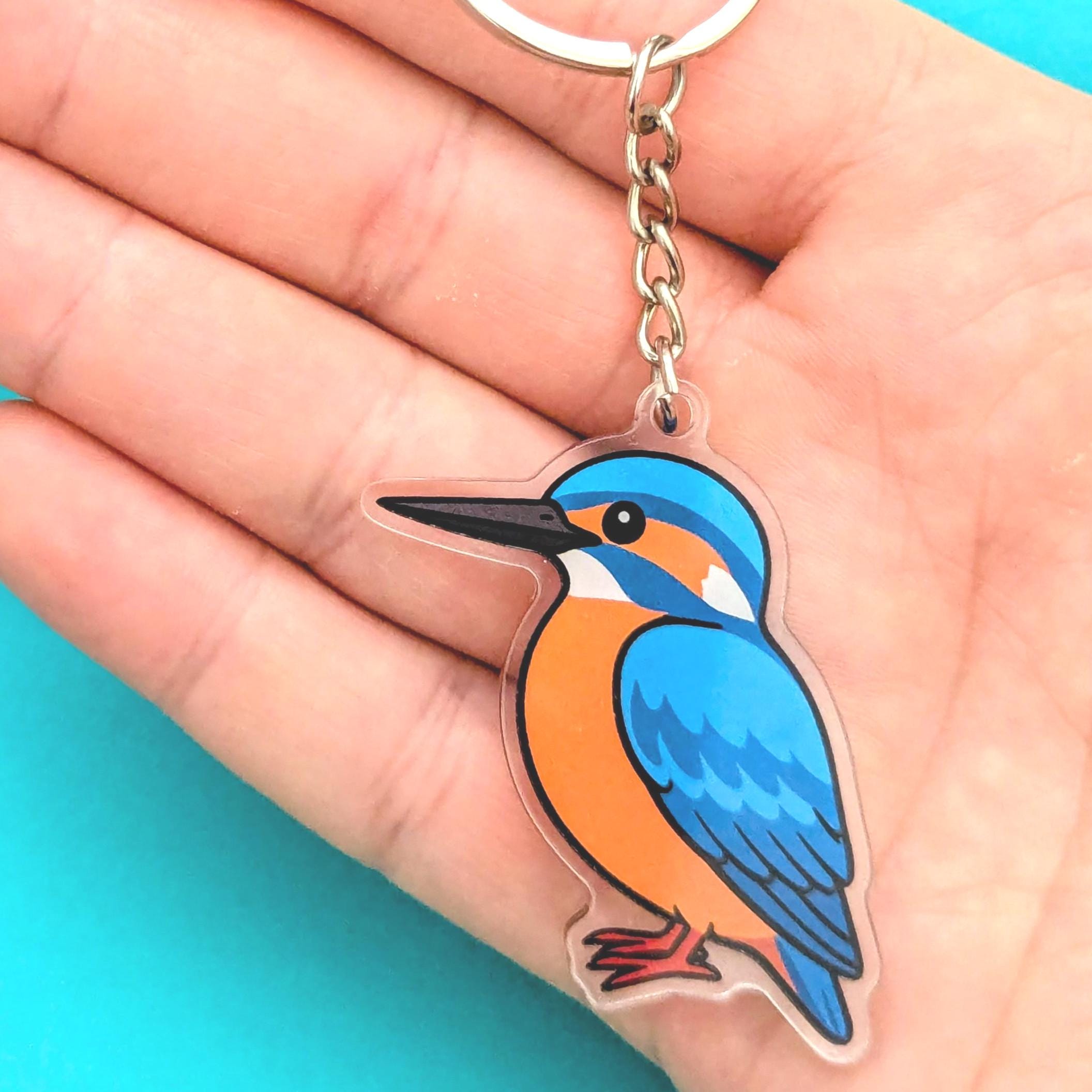 Keychain with a blue and orange bird design held in a hand against a turquoise background