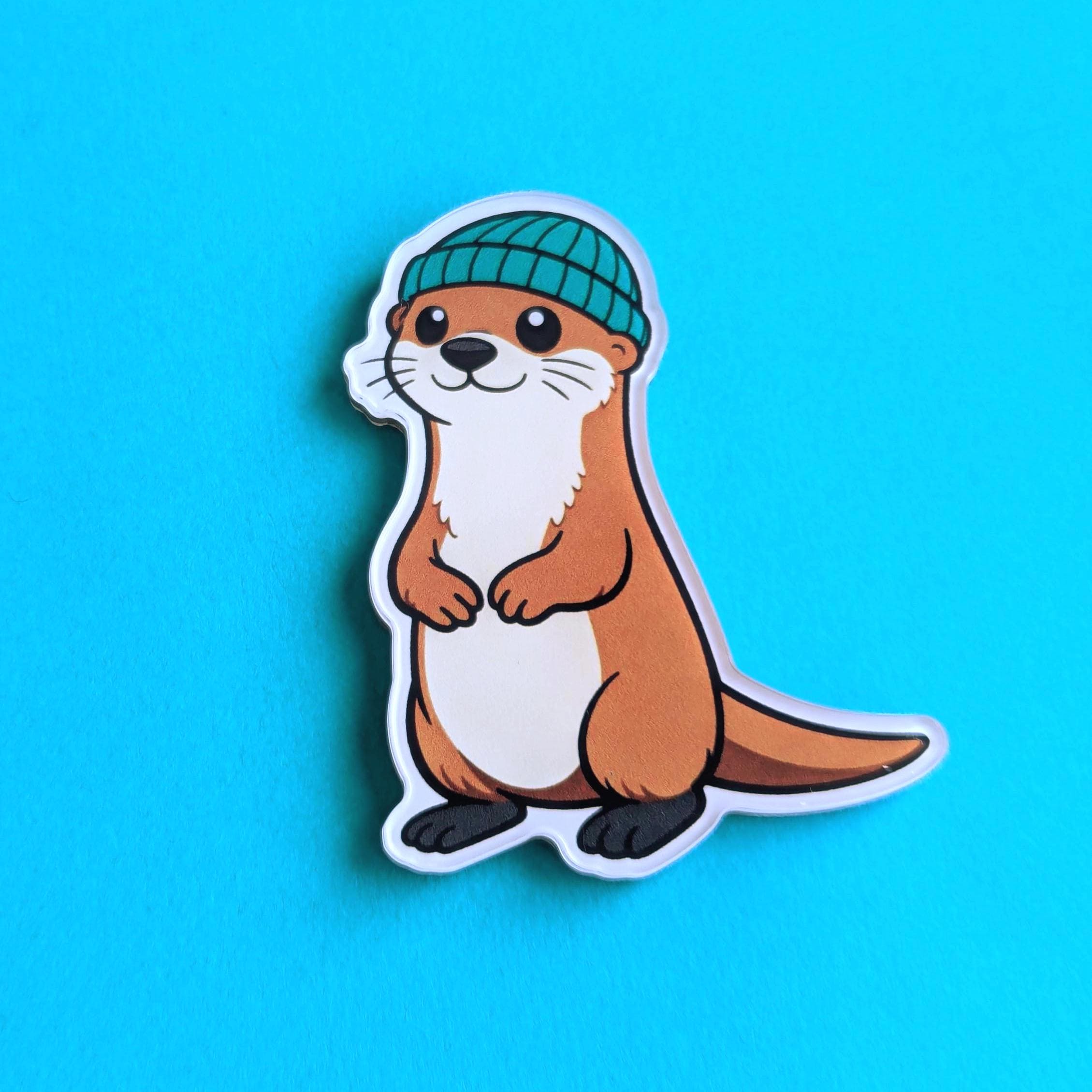 Fridge magnet of a cartoon otter wearing a green beanie on a blue background