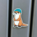 Fridge magnet of an otter wearing a blue beanie on a metallic surface