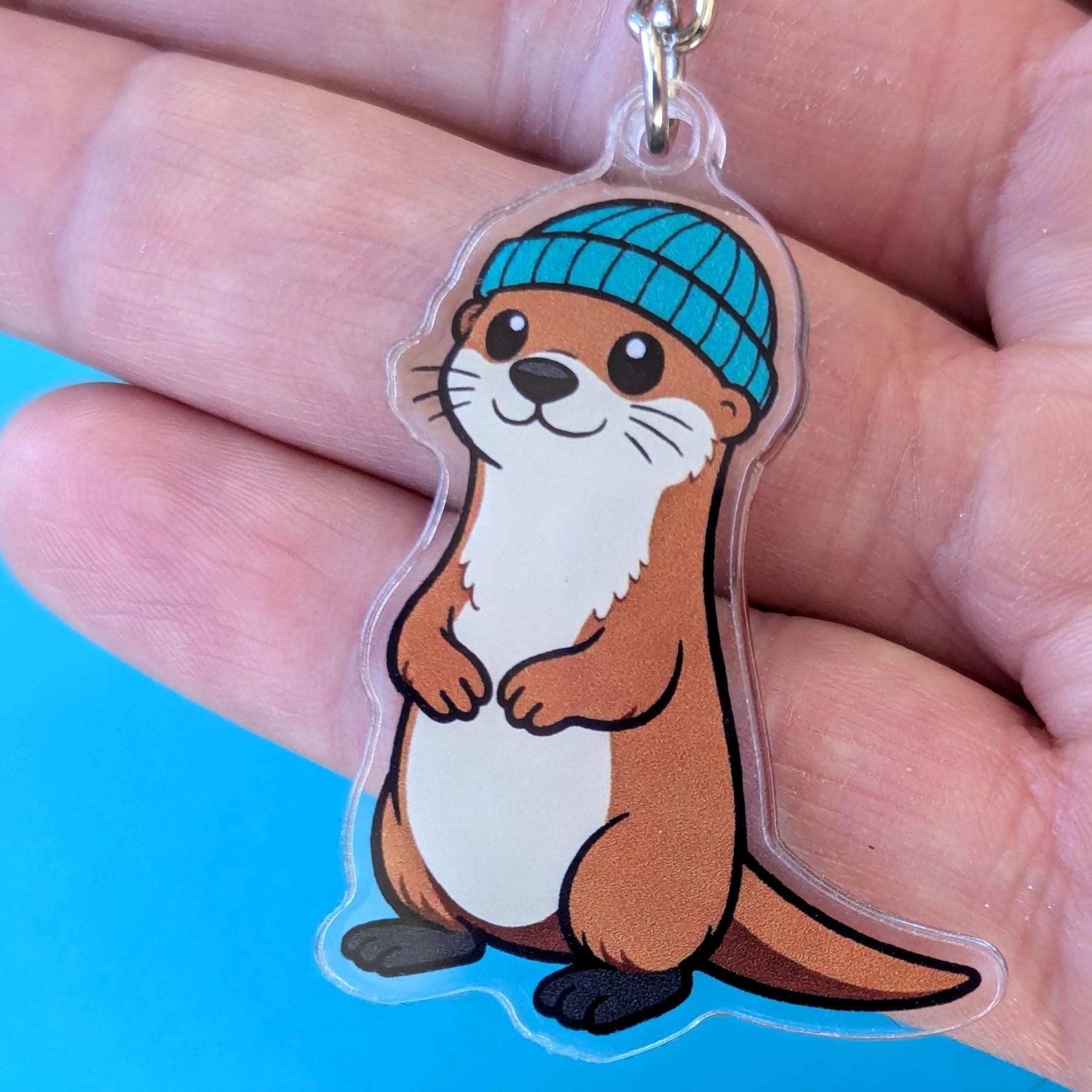 Keychain with a cartoon otter wearing a blue beanie held in a hand against a blue background