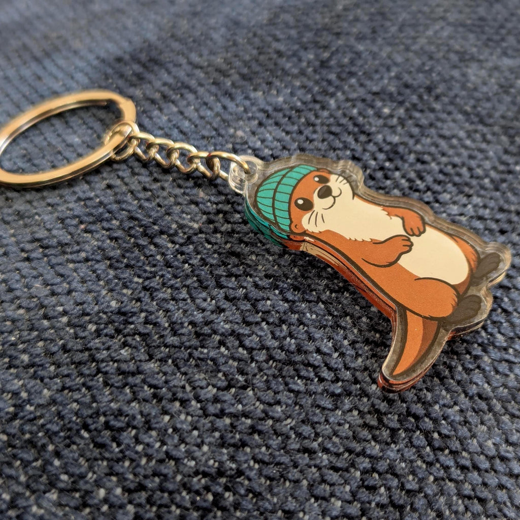 Keychain with a cartoon otter wearing a green beanie on a textured dark surface
