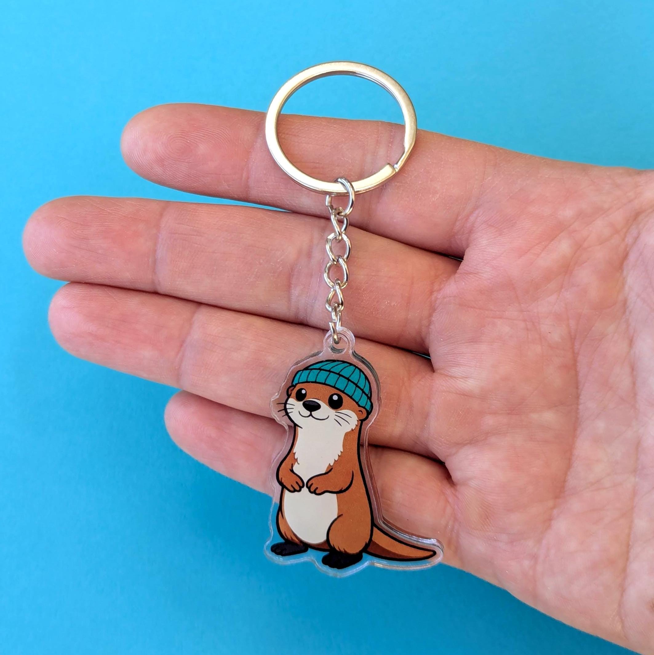 Hand holding a keychain with an otter design against a blue background