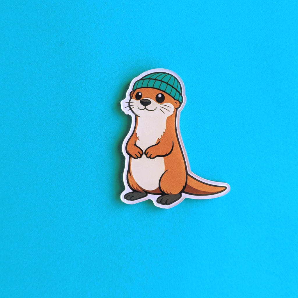 Acrylic fridge magnet of an otter wearing a green beanie on a blue background