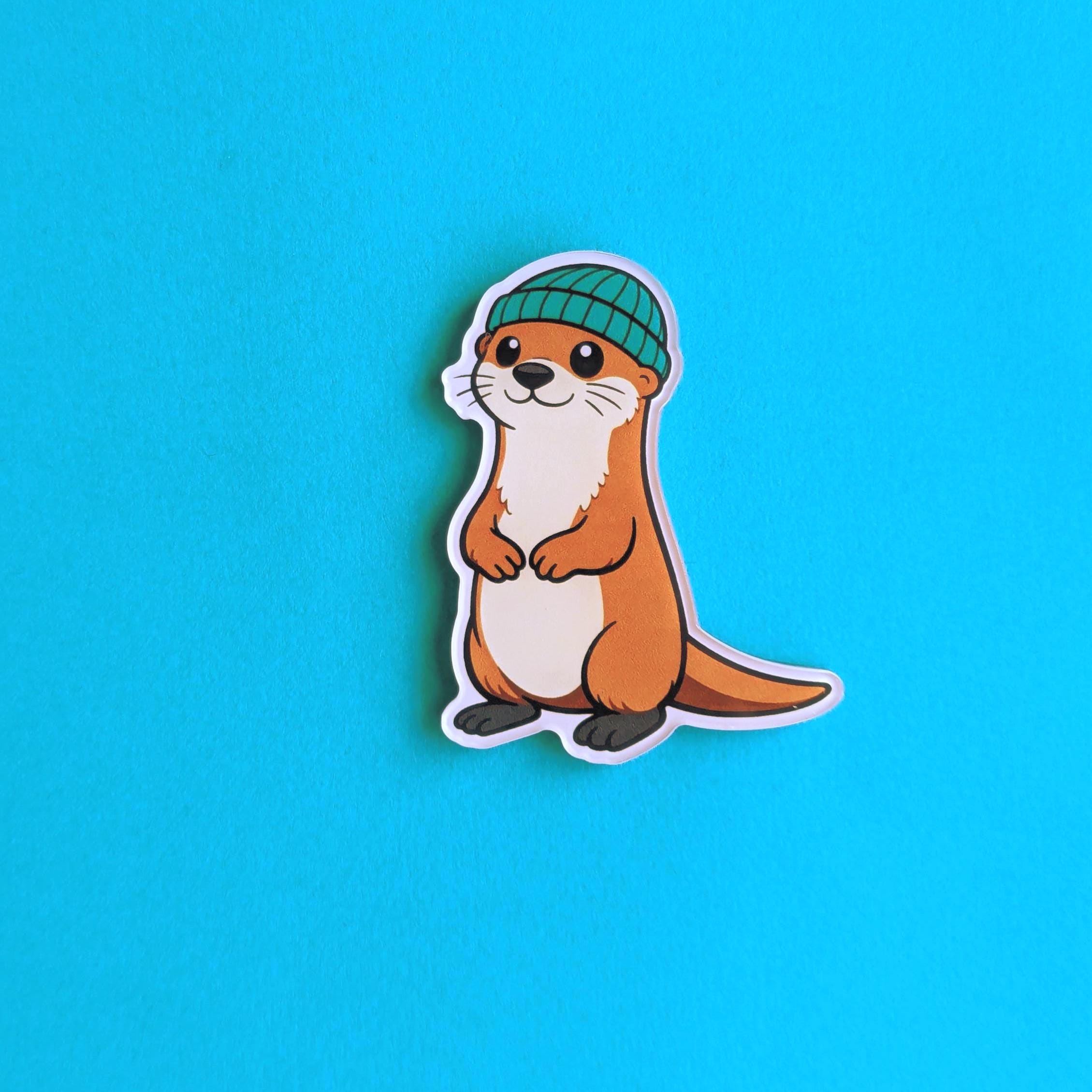 Acrylic fridge magnet of an otter wearing a green beanie on a blue background