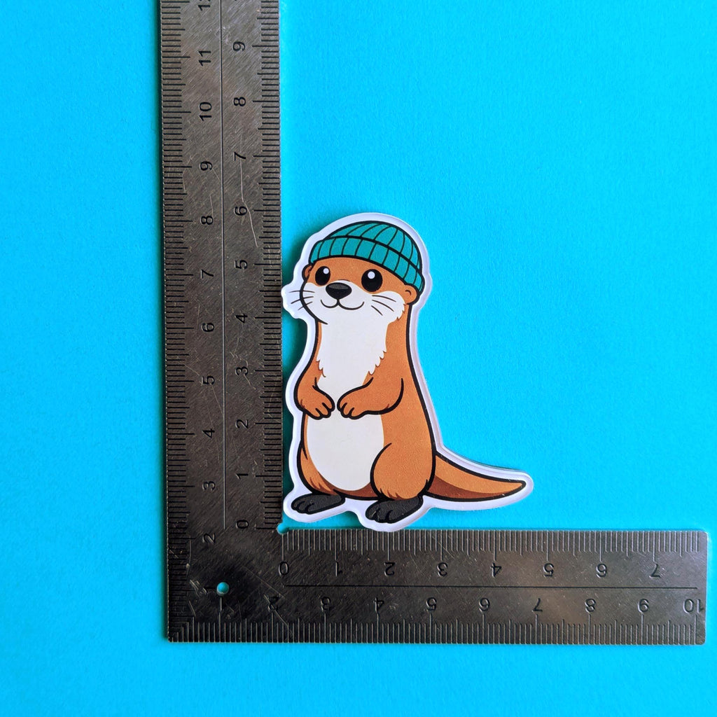 Fridge magnet of an otter wearing a beanie next to a ruler on a blue background