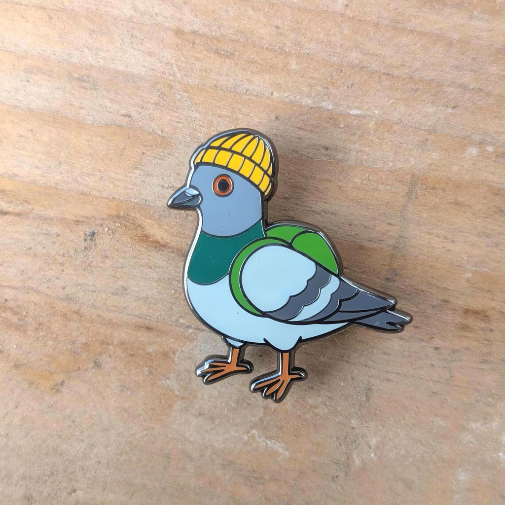 Pigeon enamel pin with a yellow hat on a wooden surface