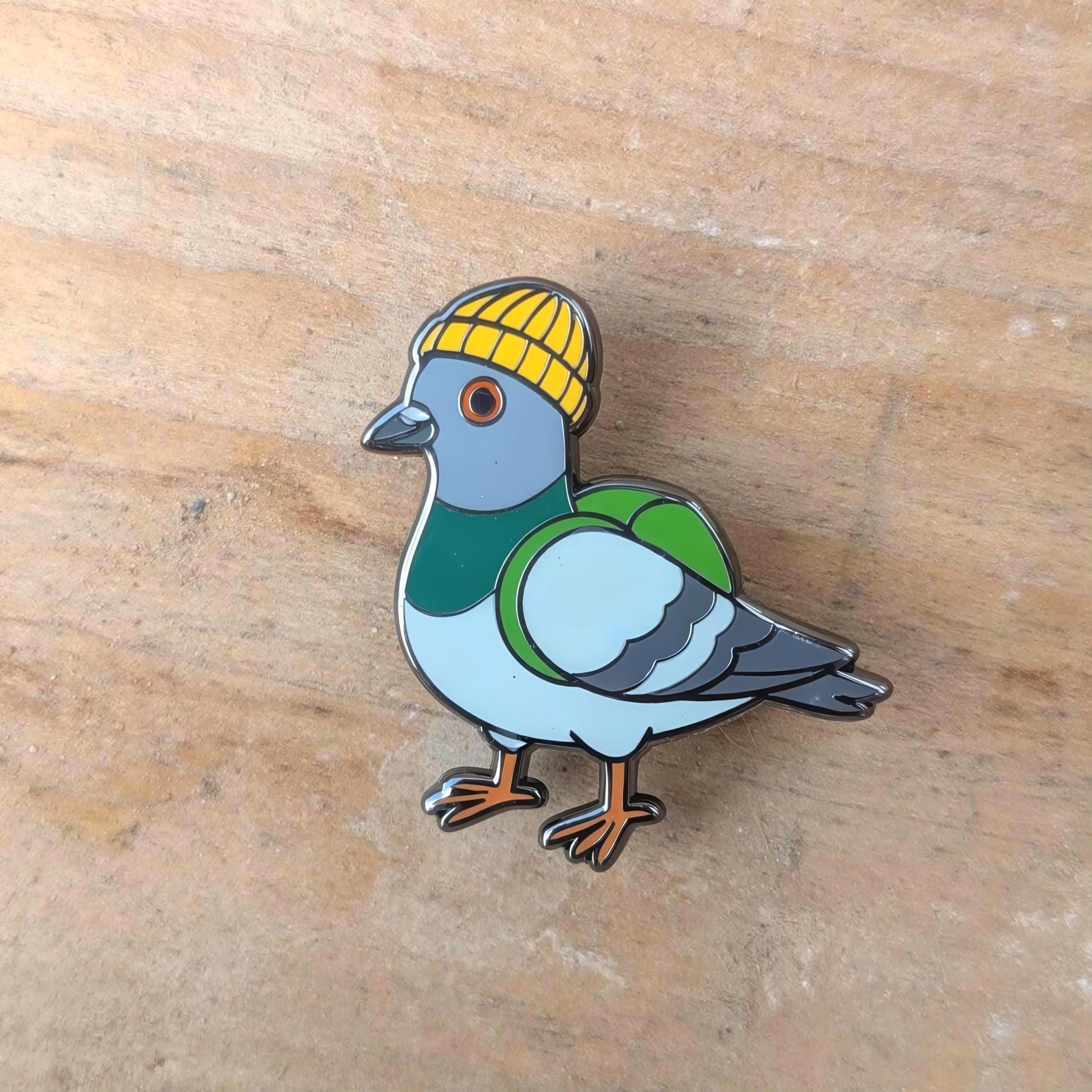 Pigeon enamel pin with a yellow hat on a wooden surface
