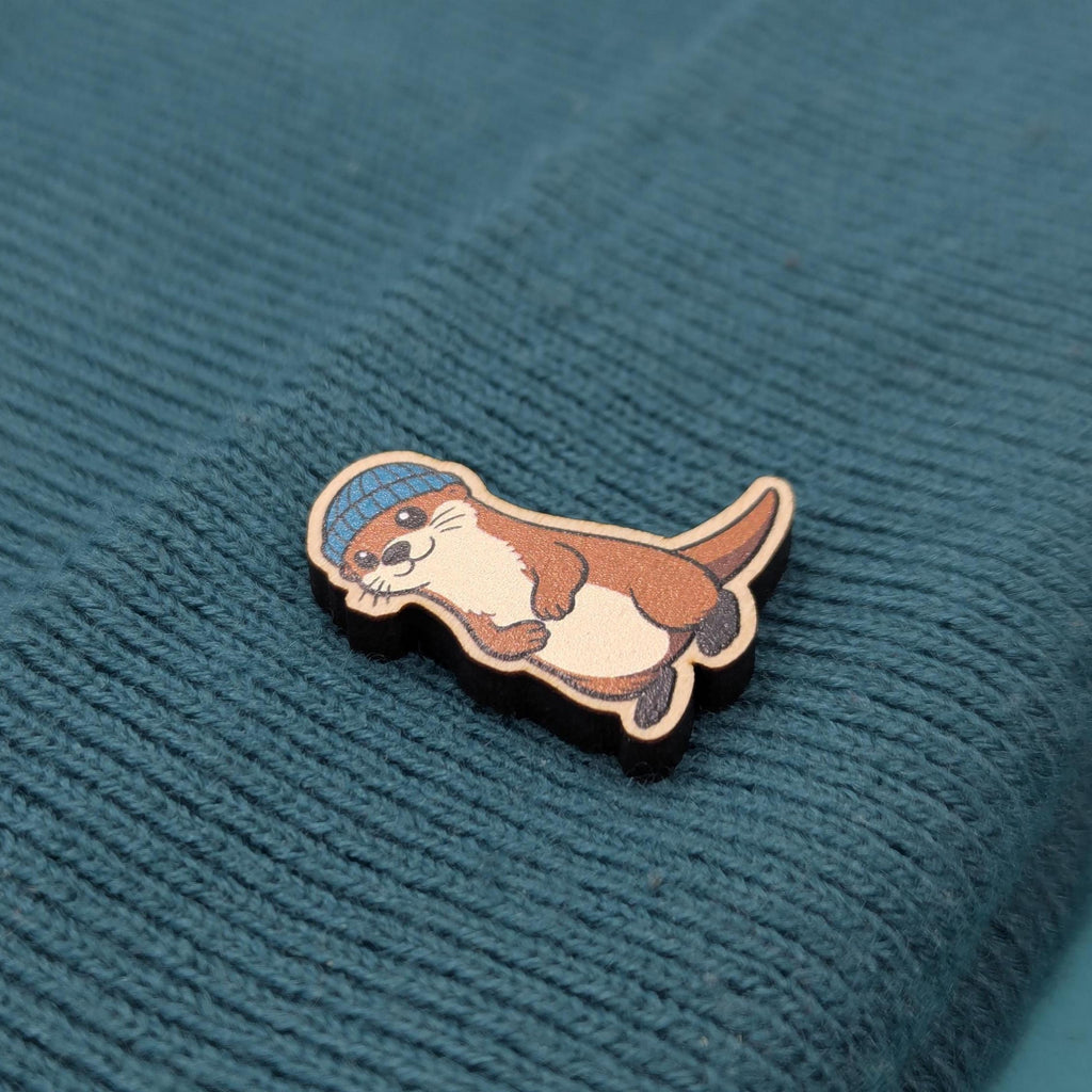 Wooden pin of an otter wearing a blue hat on a textured teal fabric background