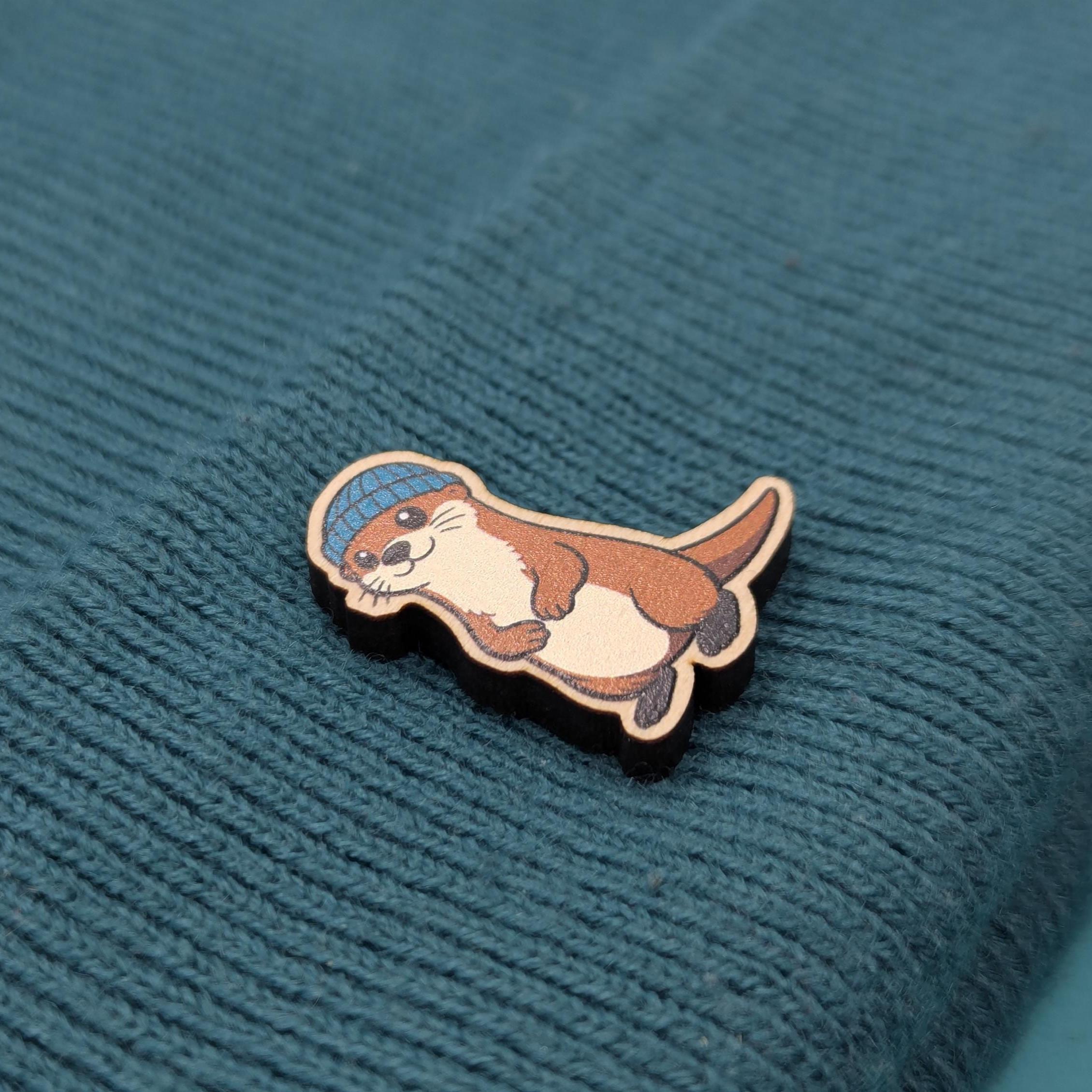 Wooden pin of an otter wearing a blue hat on a textured teal fabric background