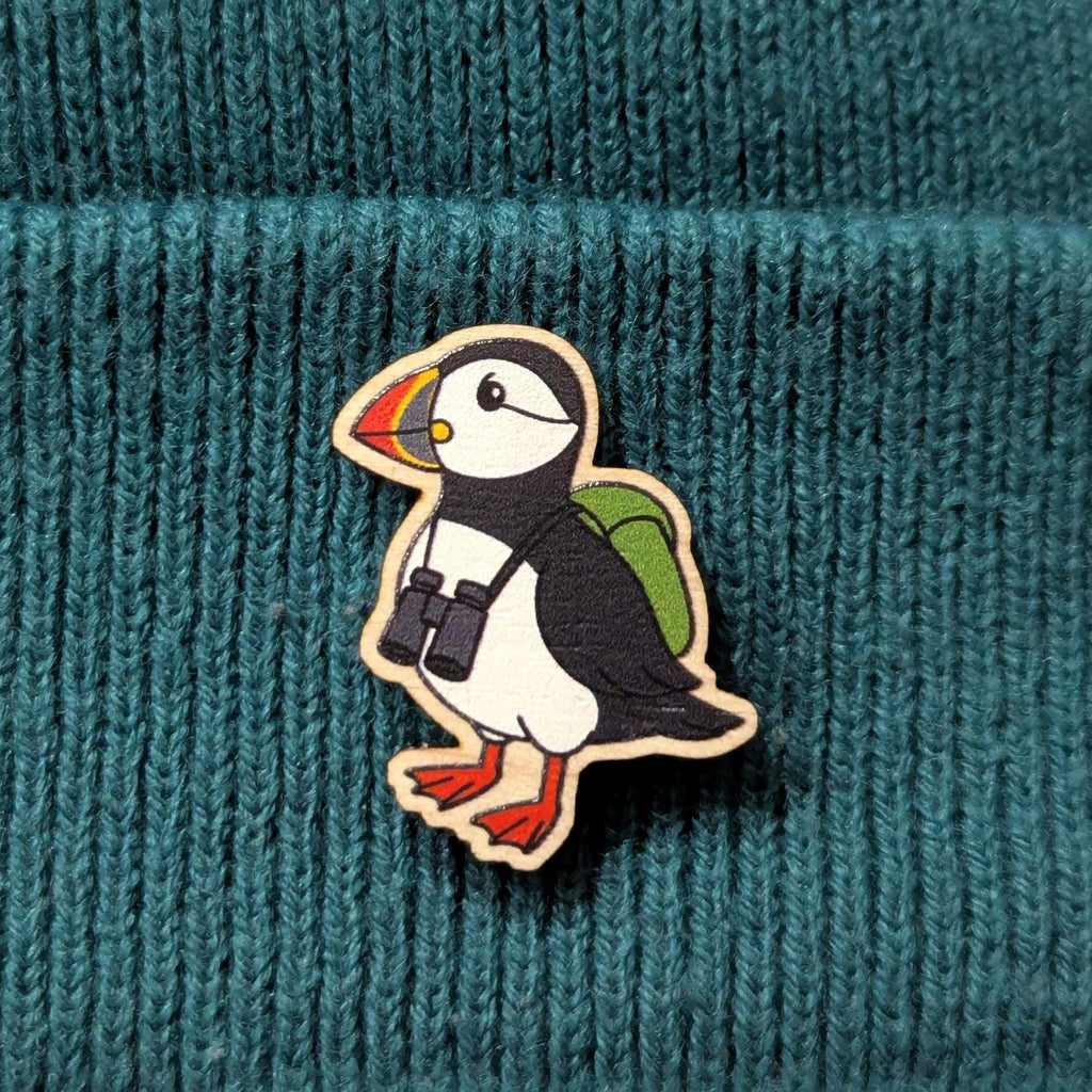Puffin wooden pin with binoculars and a backpack on a teal knitted background