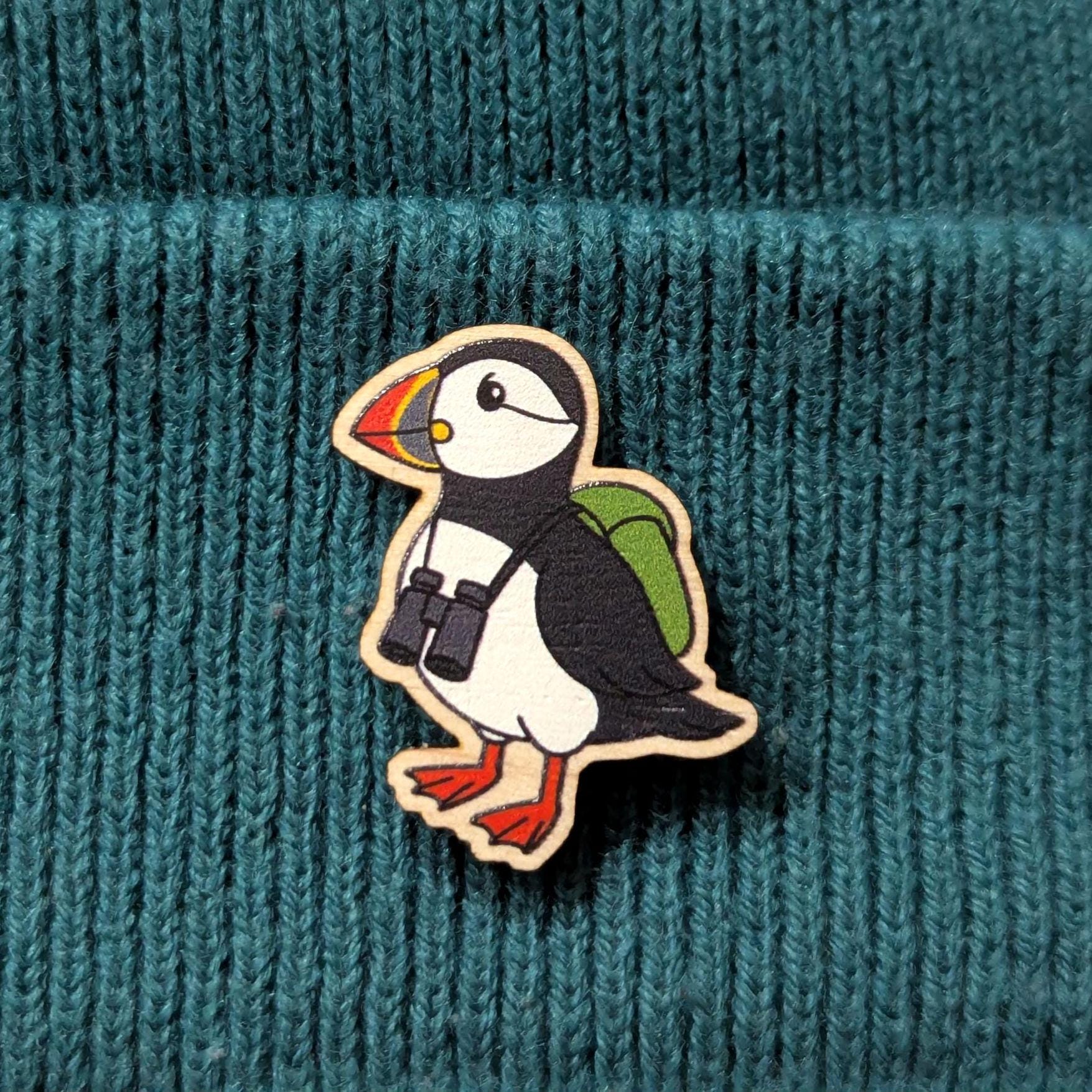 Puffin wooden pin with binoculars and a backpack on a teal knitted background