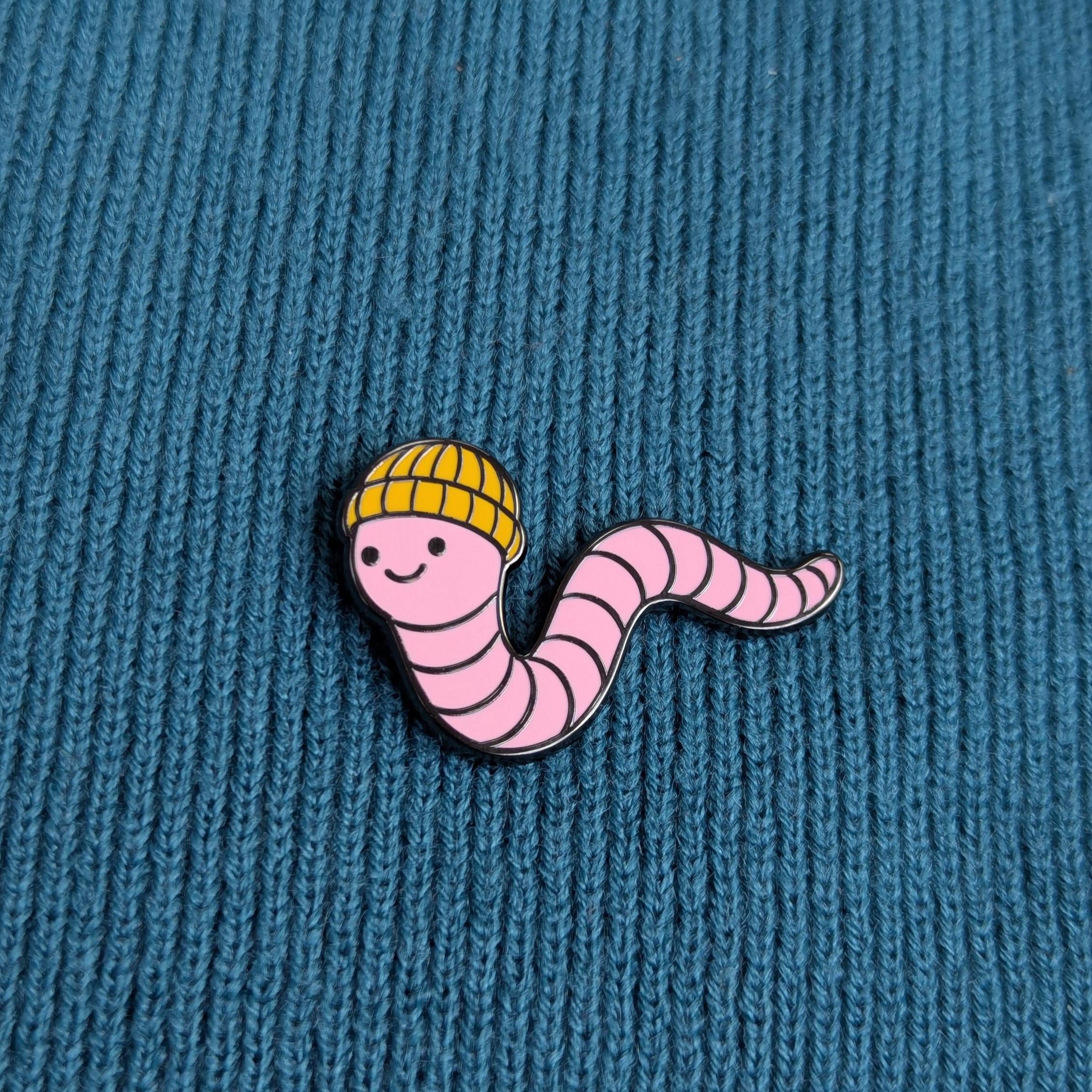 Pink cartoonish worm enamel pin badge with a yellow hat on a blue textured background