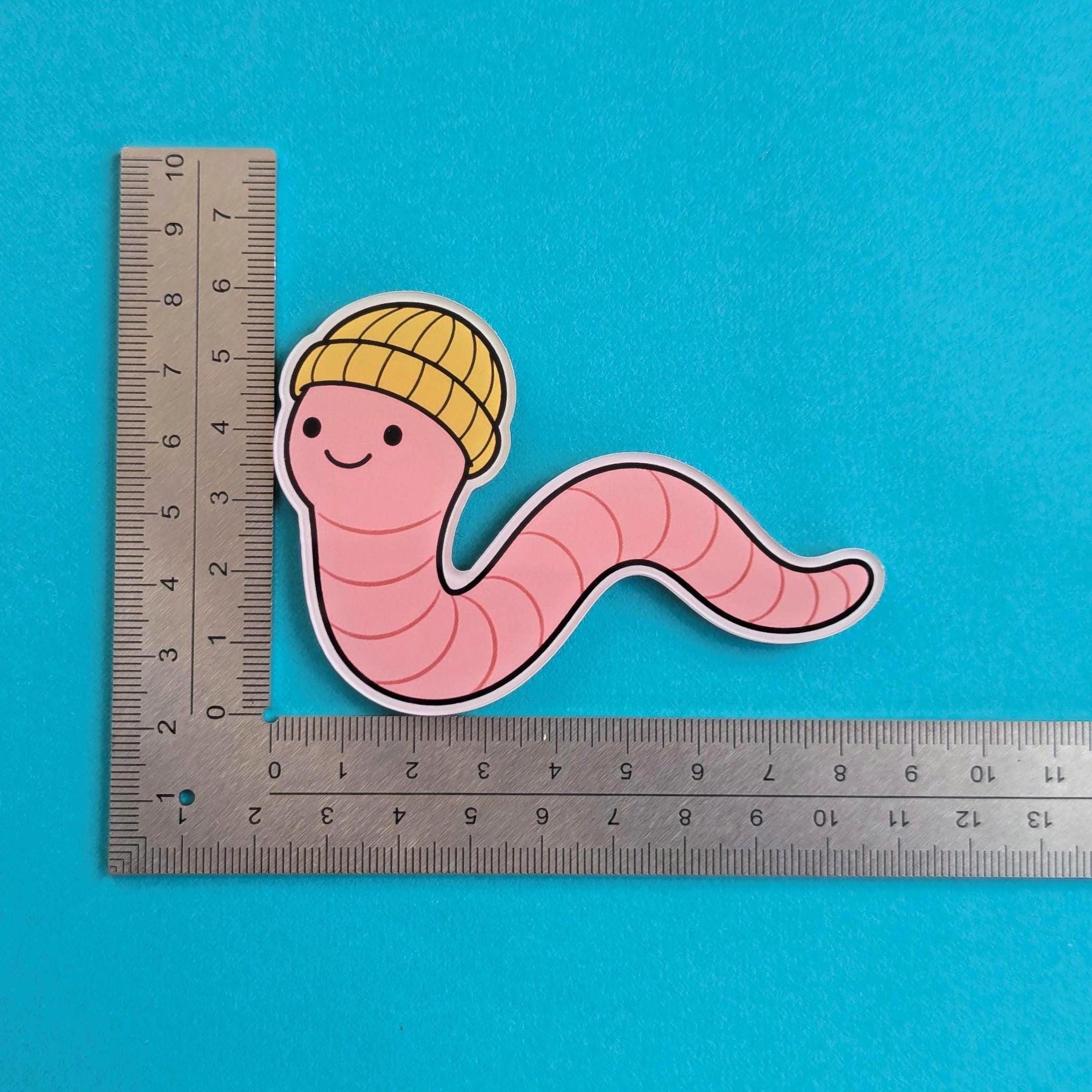 Fridge magnet of a pink worm with a yellow beanie on a blue background with a ruler for scale.
