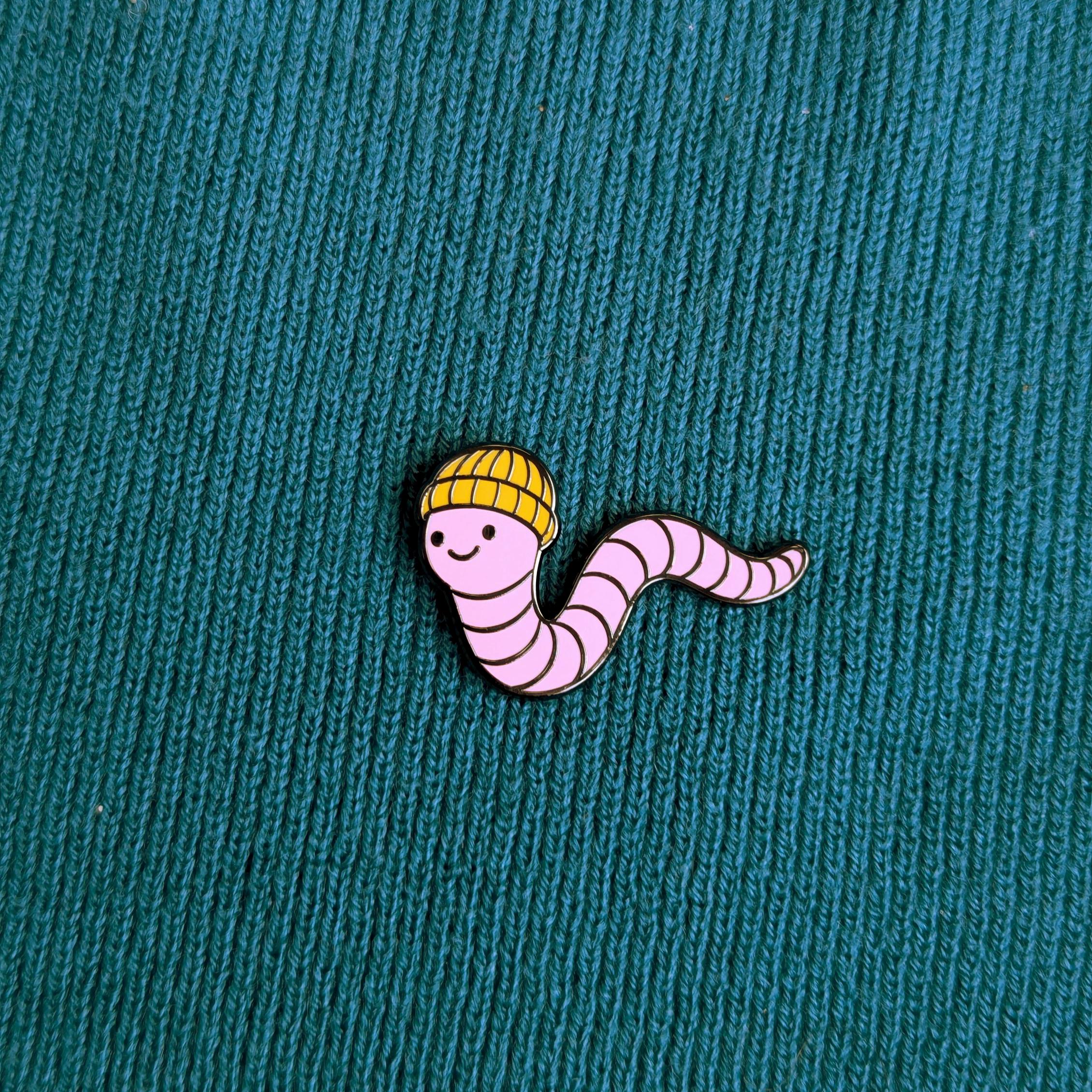 Pin of a cartoon worm wearing a yellow hat on a textured teal background
